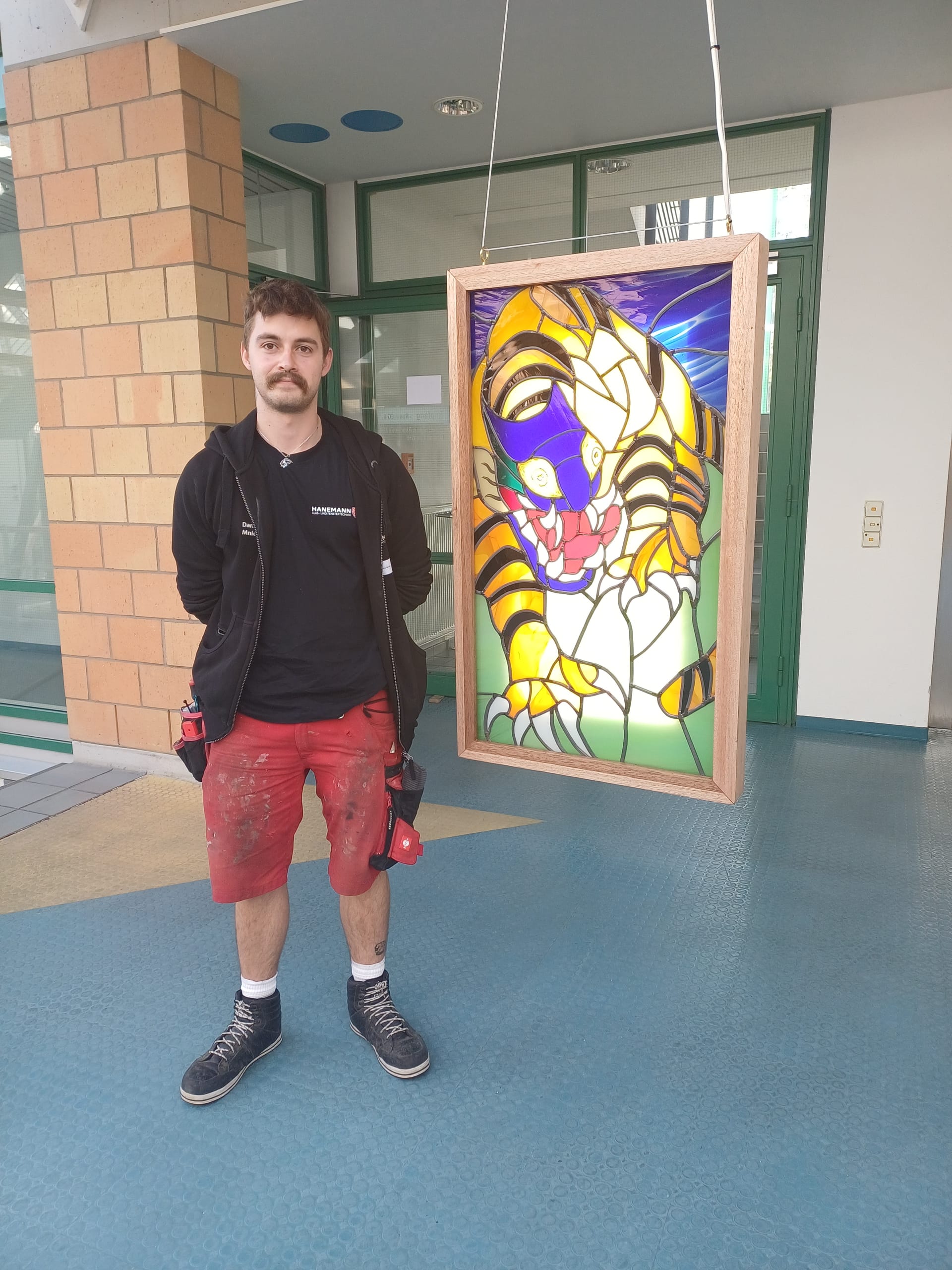 Danny Mnich von Hanemann Glas- und Fenstertechnik fertigte ein kunstvolles Bleiverglasungsgemälde, das einen fabelwesen-ähnlichen Tiger zeigt. Für das Gesicht des Tigers hat sich Mnich von japanischen Oni-Masken inspirieren lassen. Das Werk kombiniert unterschiedliche Glasarten: Der Körper des Tigers besteht aus opakem Glas, während er für den Hintergrund opales Glas verwendet hat. Hinterleuchtet mit einem LED-Panel sticht der Körper so besonders hervor. Ein Highlight sind auch die Augen, die der Meisterschüler mit Glasmalerei nochmal aufwendiger gestaltet hat.