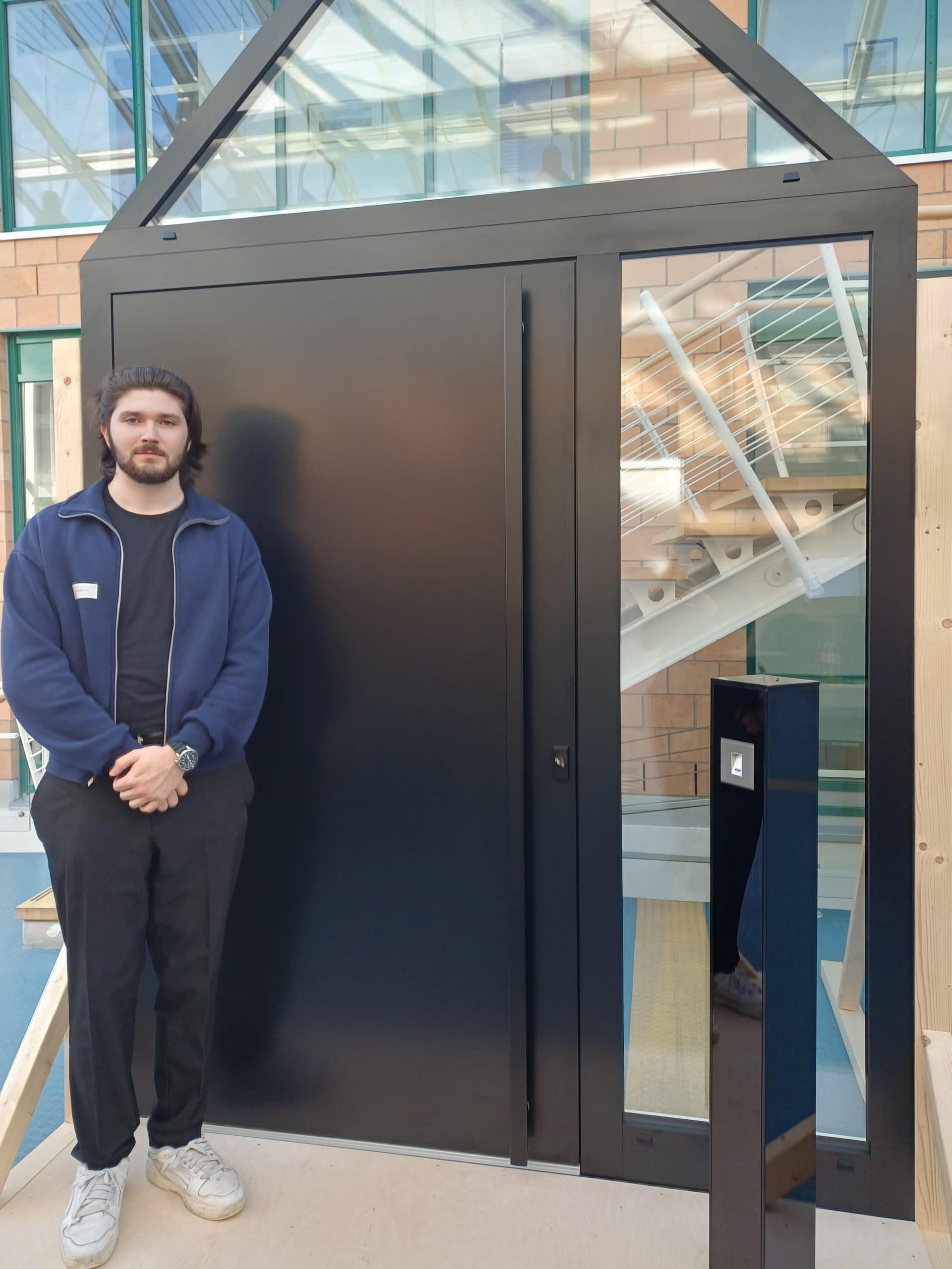 Maximilian Veil von Mundinger Fensterbau fertigte in Kooperation mit der Firma Gugelfuß eine moderne Aluminium-Haustür mit Überbreite und einer als Dreieck ausgeführten Festverglasung. Besondere Merkmale sind die verdeckt liegenden Bänder, der zwei Meter hohe Stangengriff und der freistehende Glasständer mit integriertem Fingerprint-System zur Öffnung der Tür.