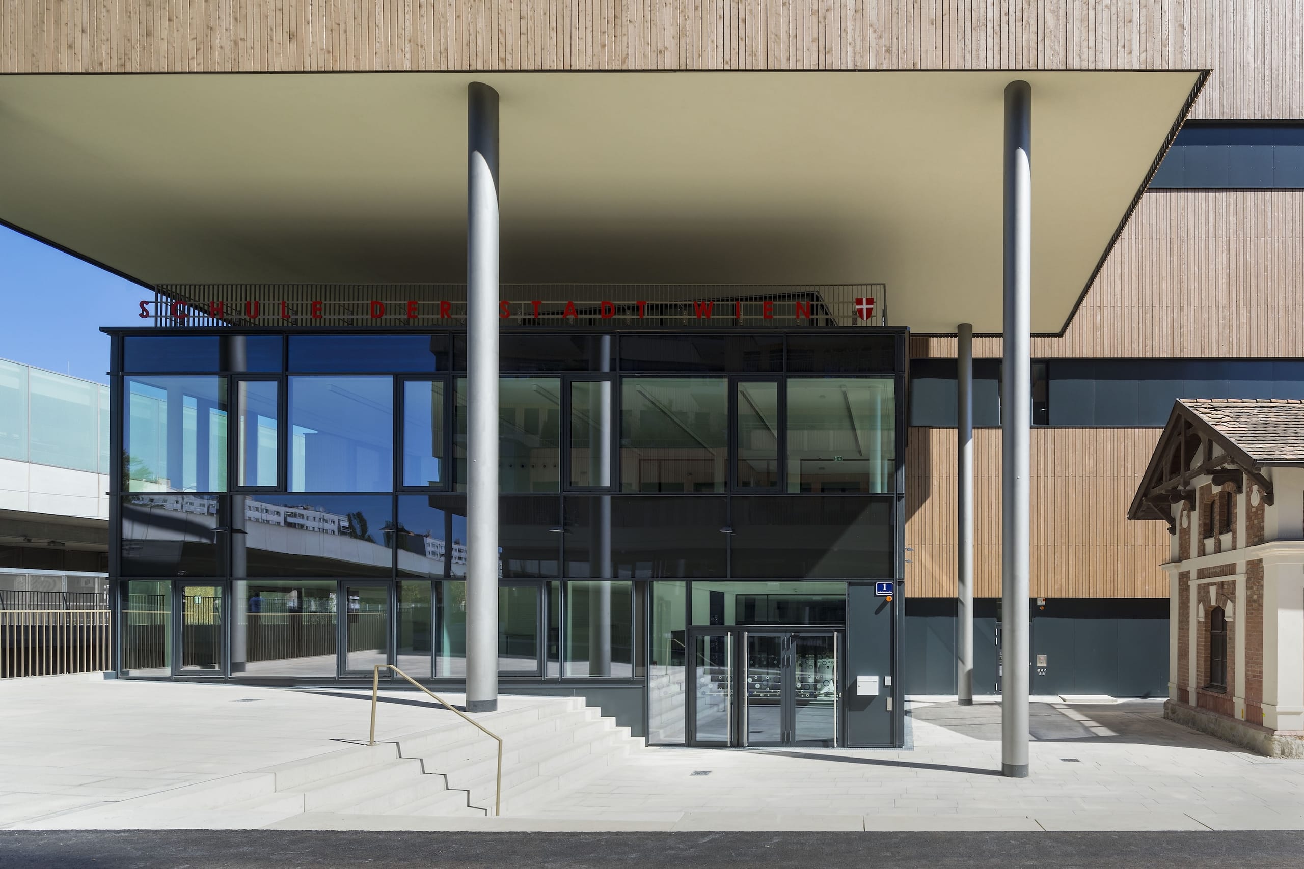 Einen Höhenunterschied im Gelände gleichen Treppen sowie unterhalb der auskragenden Gebäudeteile filigrane Stützen in unterschiedlichen Längen aus – auch dies eine gestalterische und funktionale Parallele zu den Schwestergebäuden. Am Schulhaus sind jedoch nicht nur die horizontalen Fugen deutlich betont. Tiefe vertikale Einschnitte und unterschiedlich weit hervortretende Gebäudeteile vertiefen den allgegenwärtigen Eindruck von gestapelten Quadern.